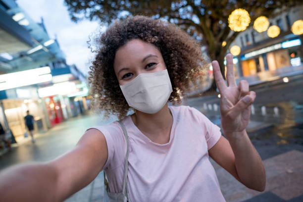 Woman wearing a facemask and taking a selfie on the street Portrait of an African American woman wearing a facemask and taking a selfie on the street making a peace sign during the COVID-19 pandemic peace sign gesture photos stock pictures, royalty-free photos & images
