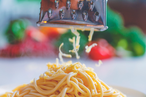 Close up of cheese being grated onto pasta.  There are vegetables out of focus in the background.