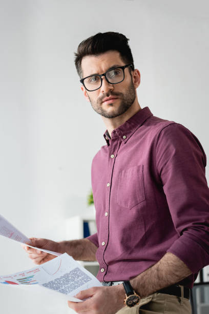 istock young, serious businessman in eyeglasses looking away while holding documents 1256210132
