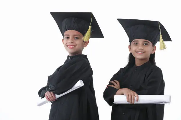 Indian, Asian Elementary age boy and girl wearing graduation caps and gowns smile while standing with degree in hand