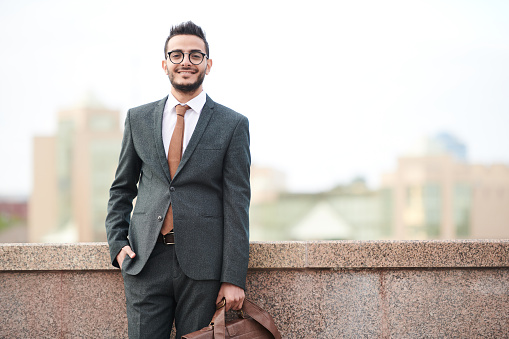 Portrait of positive handsome middle-eastern businessman with beard standing with briefcase on embankment