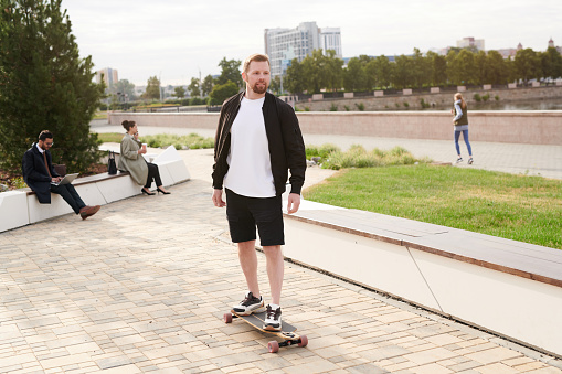 Handsome young bearded man in jacket enjoying skateboarding in city park