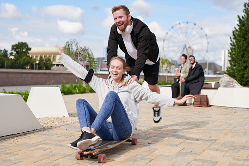 Carefree young friends riding on skateboard in city park: bearded guy pushing off ground and leaning on shoulder of girl sitting on skateboard