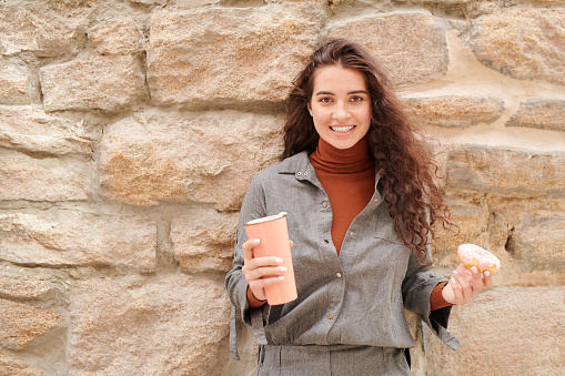 Portrait of smiling curly-haired girl in gray shirt having break for coffee and enjoying doughnut and cappuccino outdoors