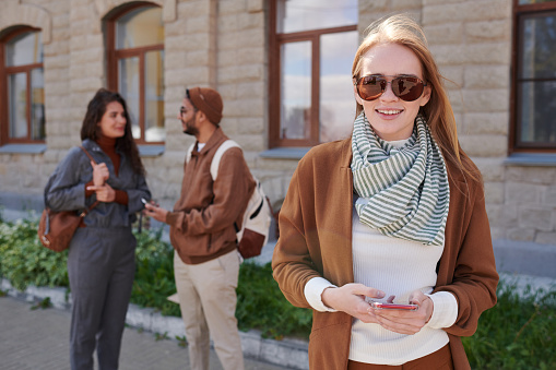 Portrait of smiling young woman in sunglasses using smartphone app outdoors while their friends chatting in background