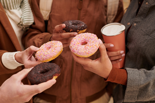 Close-up of unrecognizable friends toasting with sweet doughnuts while enjoying coffee break together