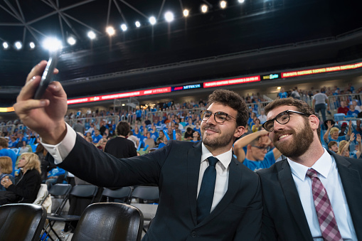 Smiling men in suit taking selfie during match in stadium.