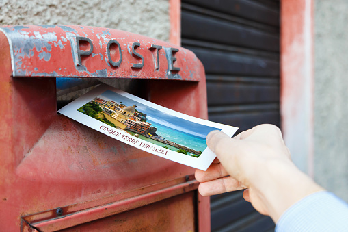 Hand drops a postcard in a red mailbox in the Vernazza, Italy. The postcard shows a view of vernazza in famous cinque terre.