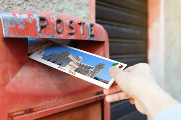 Photo of Hand drops a postcard to a red mailbox in Pisa, Italy. The postcard shows a sight of Pisa with famous tower.