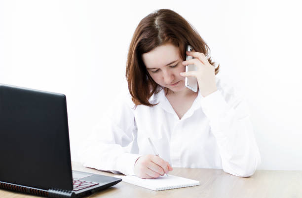 istock Portrait of student learning on line with phone and laptop taking notes in a notebook sitting at home 1256254986