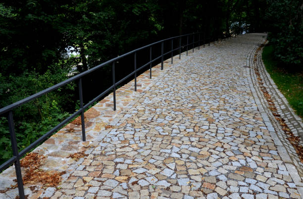 alley, row, side, down, edge, cobblestone, path, footpath, steet, irregular, cubes, pavement, paving, granite, groove, drainage, cracked, railing, metal, system, threshing, way, walkway, bike, forest, park, garden, newly, new, founded, meadow, respect, tree, trunk, preambulate, evade, evaded, channel, water, surface, gray, stone, brown, road, nature, trees, green, landscape, autumn un nuevo camino de parque hecho de cubos irregulares, plegado con un canal lateral para drenar el agua de la superficie de la carretera. piedras de granito de color gris y marrón en lugares el camino está cerrado barandilla de metal negro