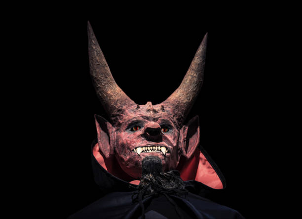 Devil with horns and in a black cloak on a dark background looks up. Man in scary mask for halloween.