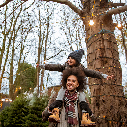 A mixed-race father and son at a Christmas tree farm together in Newcastle-Upon-Tyne. The man is carrying the boy on his shoulders, who has his arms outstretched. They are both looking away from the camera.
