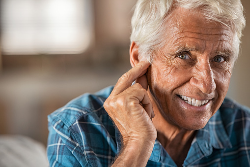 Happy man at home with hearing aid finally hears, copy space. Portrait of smiling senior man holding ear with satisfaction looking at camera. Close up face of old man relaxing at home with his new hearing aid.