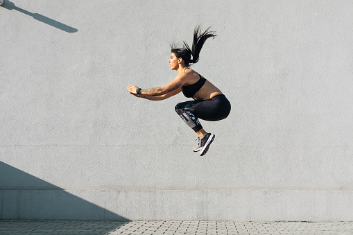 Fit young woman running and jumping in front of grey wall. Outdoor daylight wide shot.