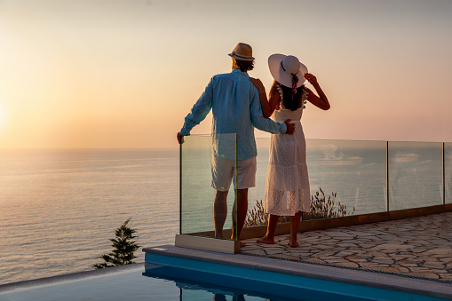 A romantic couple with hats on vacation time enjoys the summer sunset by the pool