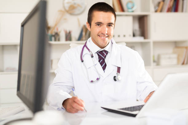 istock Positive man doctor sitting at workplace with computer 1256301561
