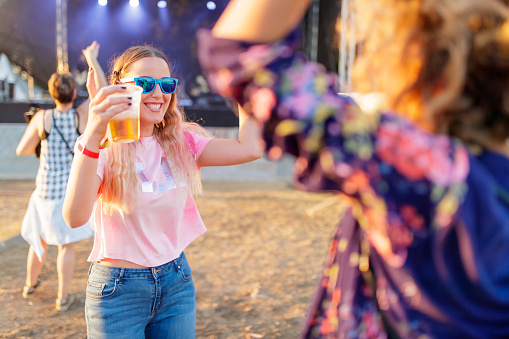 Cheerful young woman rising hands to the music and having beer on outdoor music event, blurred background for generi look