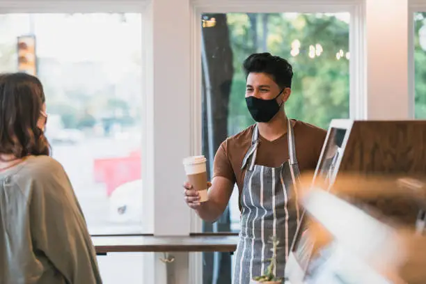 When serving his customers, the young adult male barista wears a protective mask and social distances to flatten the coronavirus spread.
