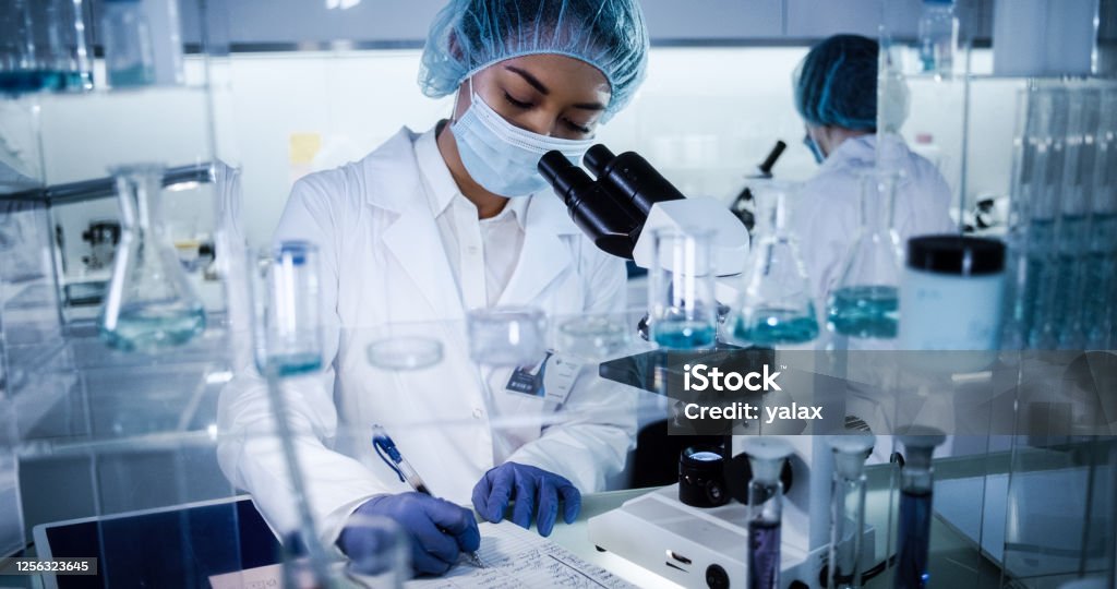 Multi ethnic, female team studying DNA mutations. Using microscope in protective workwear Scientists examines DNA models in modern Genetic Research Laboratory. Computer monitors with data in foreground Laboratory Stock Photo