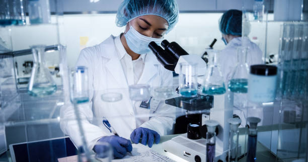 Multi ethnic, female team studying DNA mutations. Using microscope in protective workwear Scientists examines DNA models in modern Genetic Research Laboratory. Computer monitors with data in foreground science and technology stock pictures, royalty-free photos & images