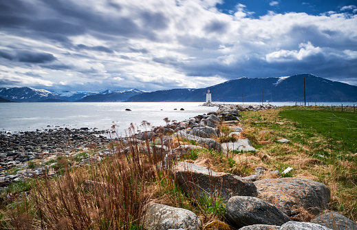 A small white lighthouse on the coastline of Norway. A path of stones leads you to it and in the foreground are many rocks as well taller and shorter grasses. The sky is filled with clouds and in the background are mountains.