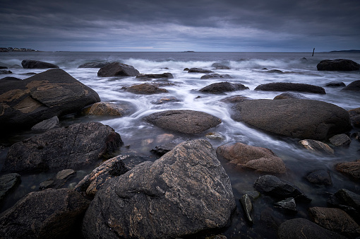 Big boulders on the coastline of Norway. Some of the rocks are under water. There are wild waves in the sea. The sky is filled with dark clouds.