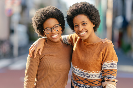 Black twin sisters are standing in the street and smiling for the camera.