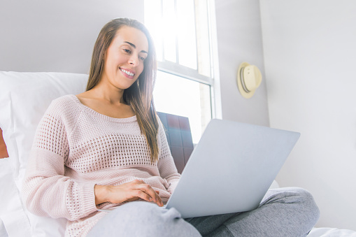 Happy Latin American woman working at home in bed using her laptop computer during the COVID-19 quarantine - new lifestyle concepts