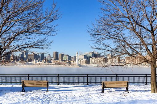 A view across the St Lawrence river to downtown Montreal. Winter shot with snow on the ground and mist rising from the frozen water. Cityscape framed by bare trees with two benches in the foreground.