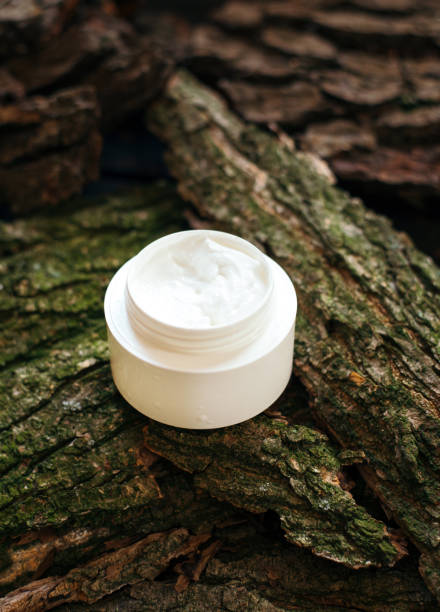 istock Cosmetic cream jar on tree bark piece side view. Organic cosmetology botanical concept. White plastic container with natural moisturizing balm and greenery. Female skin care accessory 1256350546