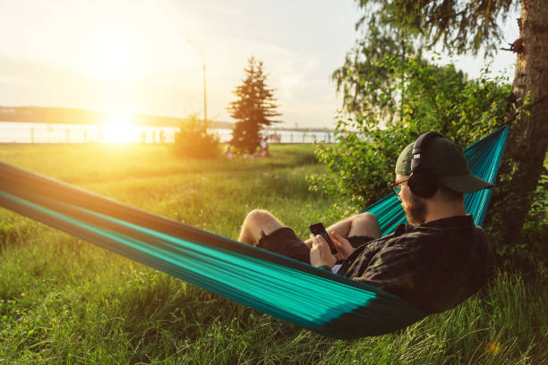 hipster mâle moderne retenant le smartphone, se relaxant et appréciant écouter la musique du service de streaming dans le hamac dans le parc au coucher du soleil - hamac telephone homme photos et images de collection
