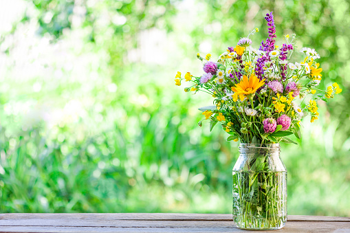 Wildflowers in glass jar stand on wooden table in the garden. Summer rural scene. Copy space.