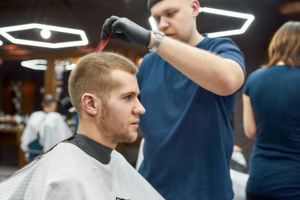 istock Professional tattooed barber making modern haircut for a young handsome guy sitting sitting barbershop chair. Focus on client 1256369612