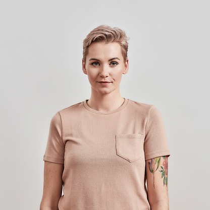 Portrait of a young attractive tattooed woman with pierced nose and short hair in beige t shirt looking at camera, standing isolated over light background. Front view