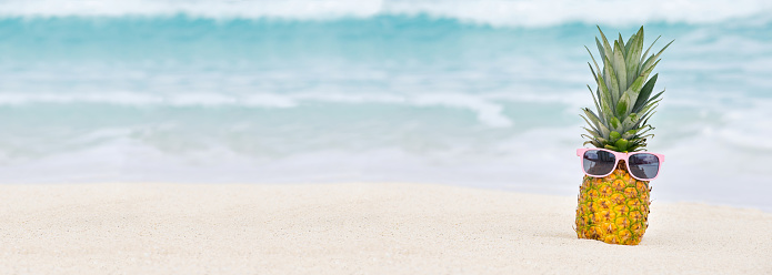 pineapple fruit with sunglasses and flip flop sandals on the beach sand against a blue sky with clouds, summer vacation concept, copy space, selected focus