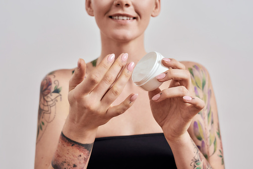 Cropped portrait of smiling tattooed woman trying skin care product, holding white plastic jar of cream or body lotion isolated over grey background. Selective focus on hands and jar. Front view