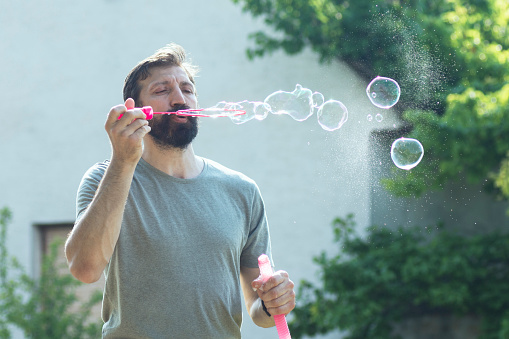 The bearded man is blowing soap bubbles outdoors.