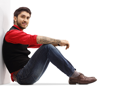 Young man in a red shirt and jeans sitting and leaning on a wall isolated on white background