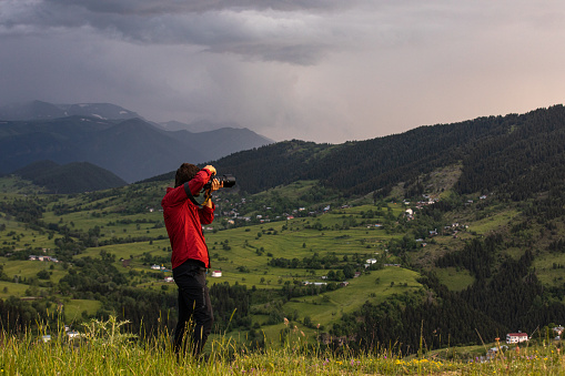 Shot of a young male traveler photographer with red shirt looking at mountain scenery with camera and taking beautiful spectacular photos during sunset in famous Şavşat Town, Artvin in Black sea region of Turkey on sunny summer day