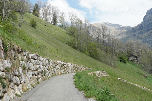 A narrow asphalt road build in the slope of an Alpine hill, reinforced by a stone wall to prevent land sliding. The steep slope is covered with fresh green grass in the springtime. Region Engelberg, canton Obwalden in Switzerland