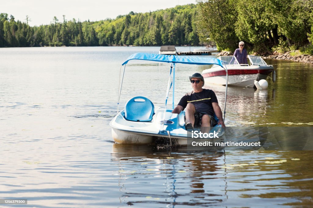 50+ couple enjoying vacations on nautical vessel. 50+ couple enjoying vacations and a sunny day. Man is on a pedal boat, woman on a small motor boat on a lake. They are wearing casual clothes and sunglasses. Horizontal full length outdoors shot with copy space. Pedal Boat Stock Photo
