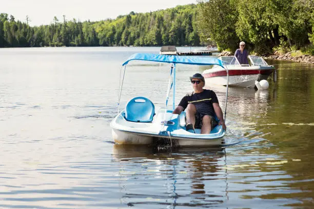 50+ couple enjoying vacations and a sunny day. Man is on a pedal boat, woman on a small motor boat on a lake. They are wearing casual clothes and sunglasses. Horizontal full length outdoors shot with copy space.