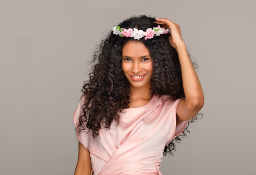 Pretty young Dominican bridesmaid in a pink dress holding a coronet of flowers to her long curly hair with a happy charming smile against a beige studio background