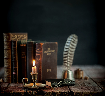 Vintage study area with books, quill pen and candle on an old wood desk against a black background.