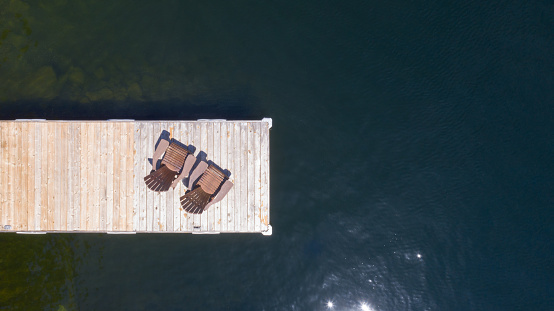 Drone aerial view of Two Adirondack chairs on a wooden pier overlooking the calm water of a lake in Muskoka, Ontario Canada.