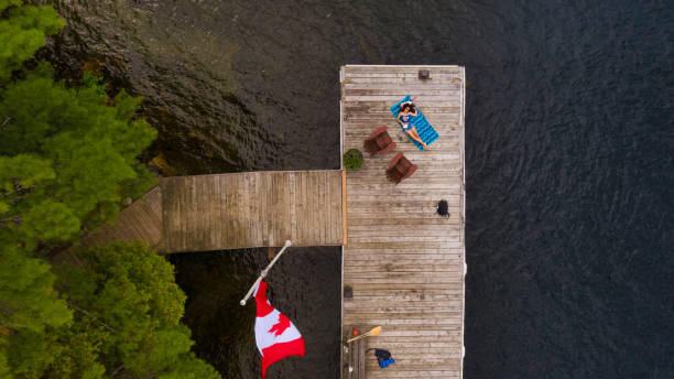 Aerial view of a wooden pier in a lake in Muskoka with a young woman tanning Aerial view of a wooden pier in a lake in Muskoka with a young woman tanning on a beach towel and two Adirondack chairs. The Canadian flag is flying on the pole. cottage life stock pictures, royalty-free photos & images