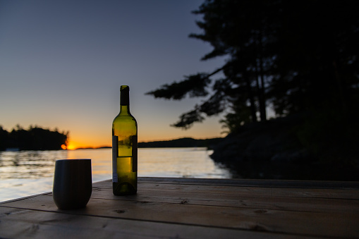 Bottle of wine and a tumbler glass on a cottage wooden pier facing the calm water of a lake during summer sunset.