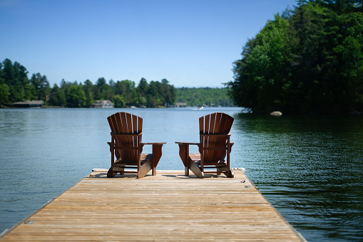 Two Adirondack chairs sitting on a wooden pier facing the calm water of a lake in Muskoka, Ontario Canada. A cottage nestled between trees is visible in background.