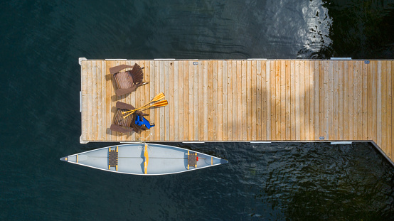 Drone aerial view of two Adirondack chairs on a wooden pier facing the blue water of a lake in Ontario. A yellow canoe is tied to the dock. Life jacket and oars are visible near the chairs.